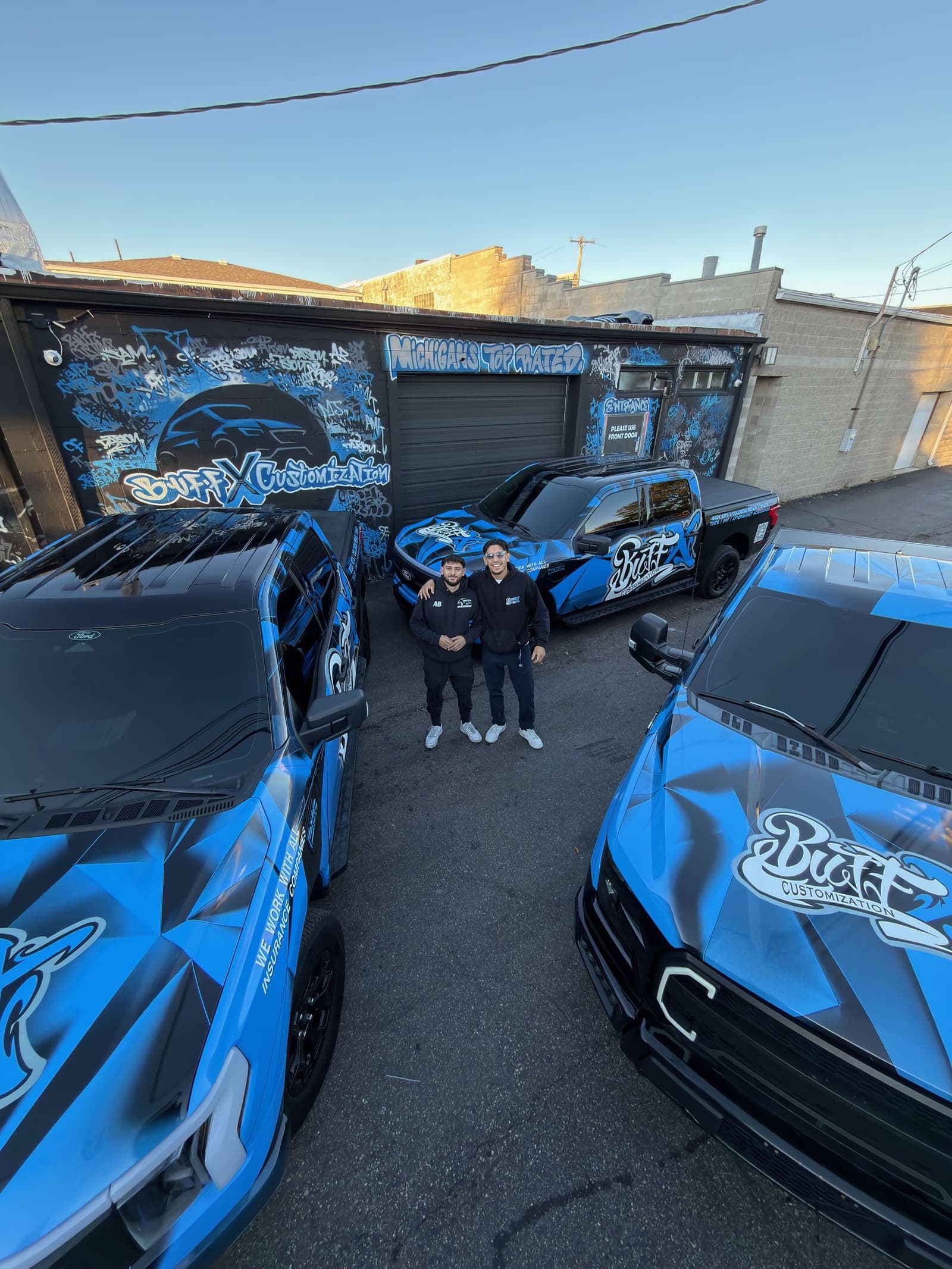 Co-founders Abbas Khattab and Sam Alsaadawi outside the Buff X Customization shop on W Warren St, between three Buff X wrapped trucks
