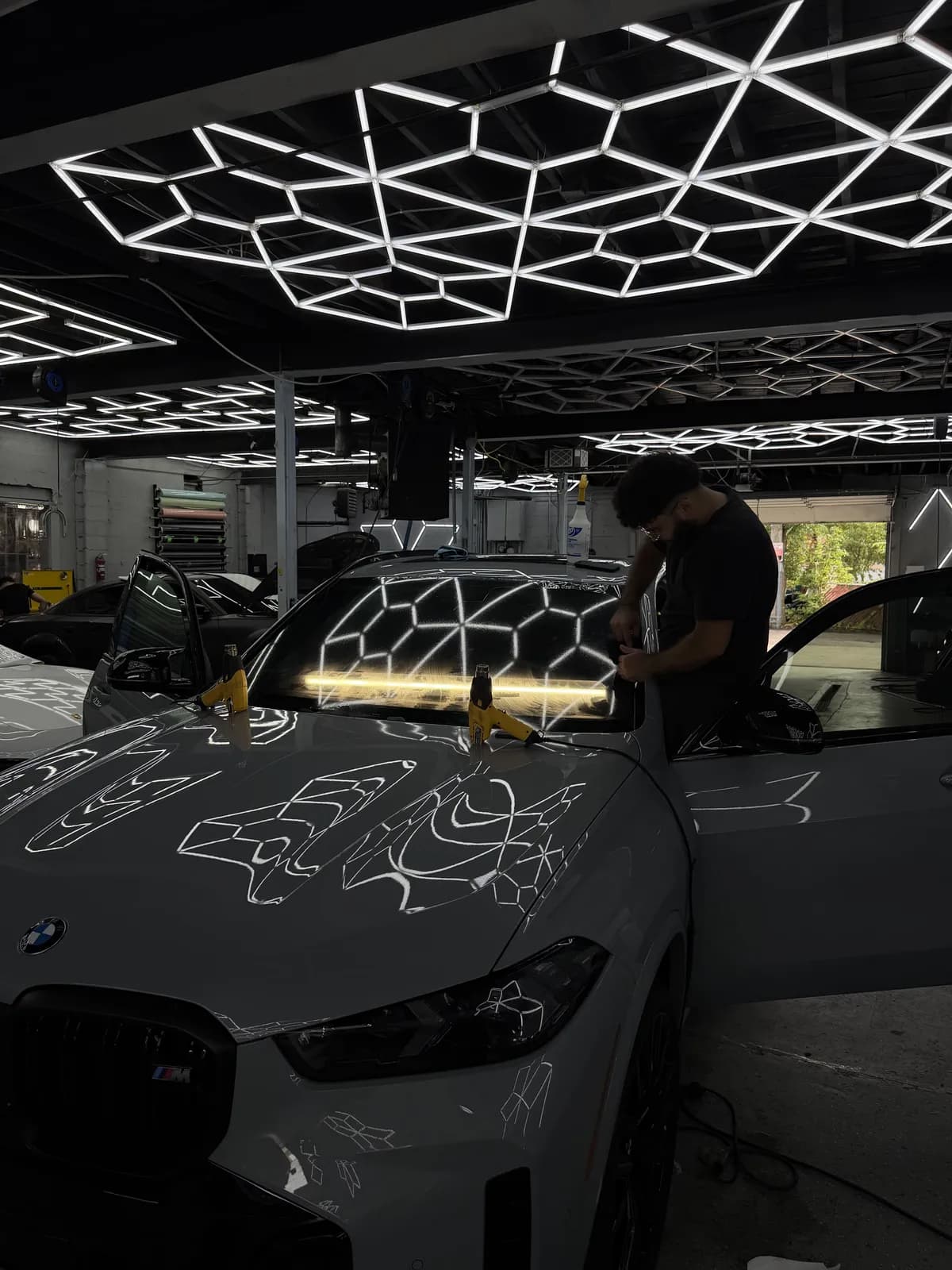 Installer fitting windshield protection film on a gray BMW X5 M under the shop lights at Buff X Customization in Dearborn Heights